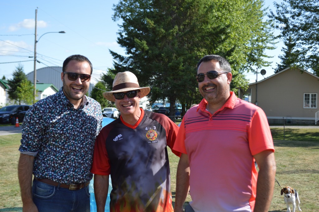 Trois hommes souriants posant côte à côte dans un parc, dont un portant un chandail du service d’incendie de Fassett, par une journée ensoleillée.