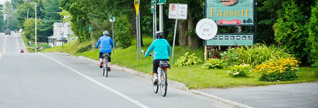 Deux cyclistes circulent sur la route près du panneau d’accueil « Fassett – Bienvenue ».
