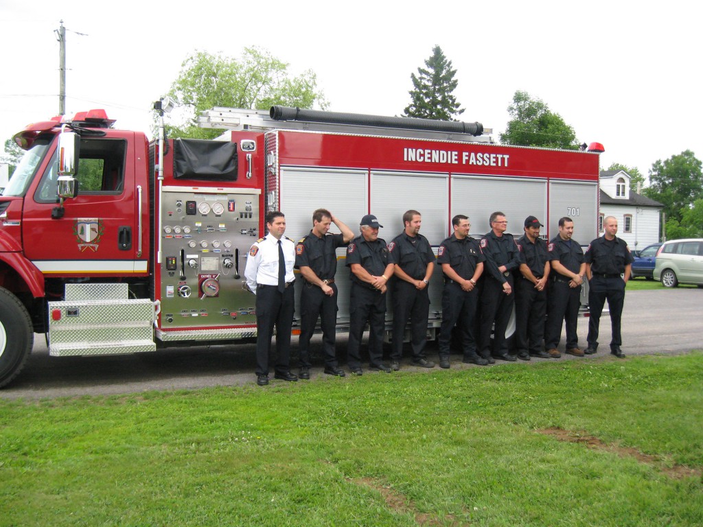Neuf pompiers du service d’incendie de Fassett alignés devant un camion d’incendie rouge portant l’inscription « Incendie Fassett ».